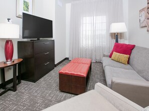 Ein Hotelzimmer mit Flachbildfernseher auf einem Schrank, einem roten Hocker, einem grauen Sofa mit rotem und gelbem Kissen und einer weißen Lampe.