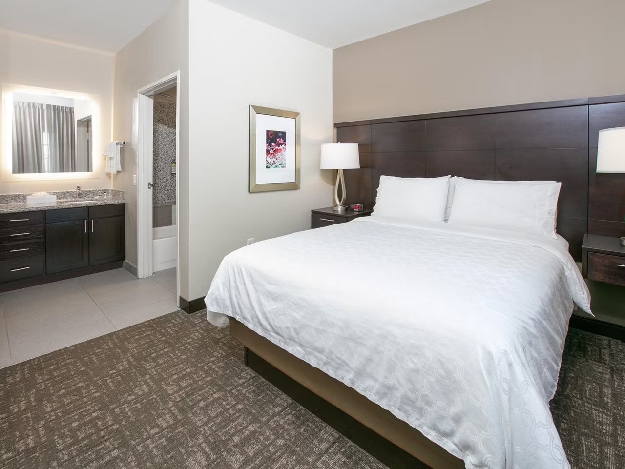 A hotel room with a bed, bedside table, lamp, and a bathroom with a sink and mirror.