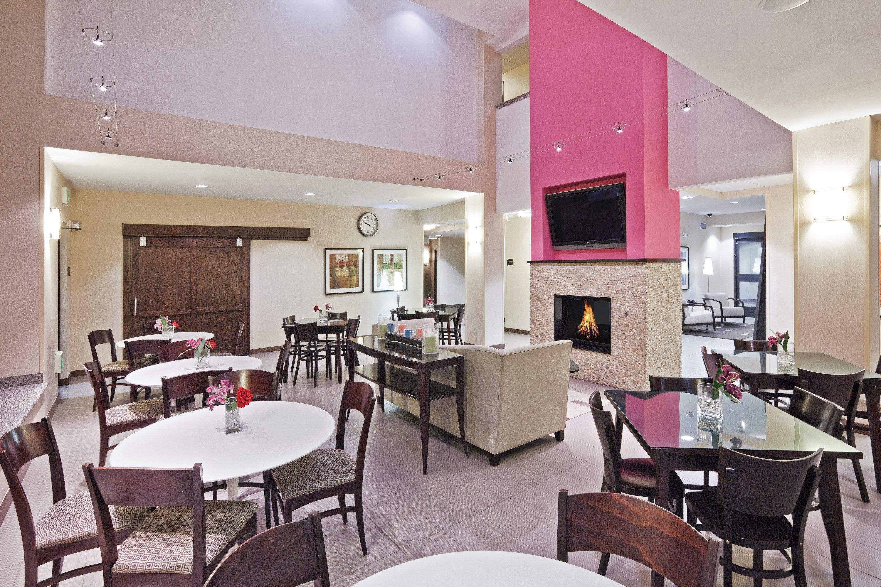A spacious dining area with round tables and chairs, a fireplace, and a television mounted on a pink wall.