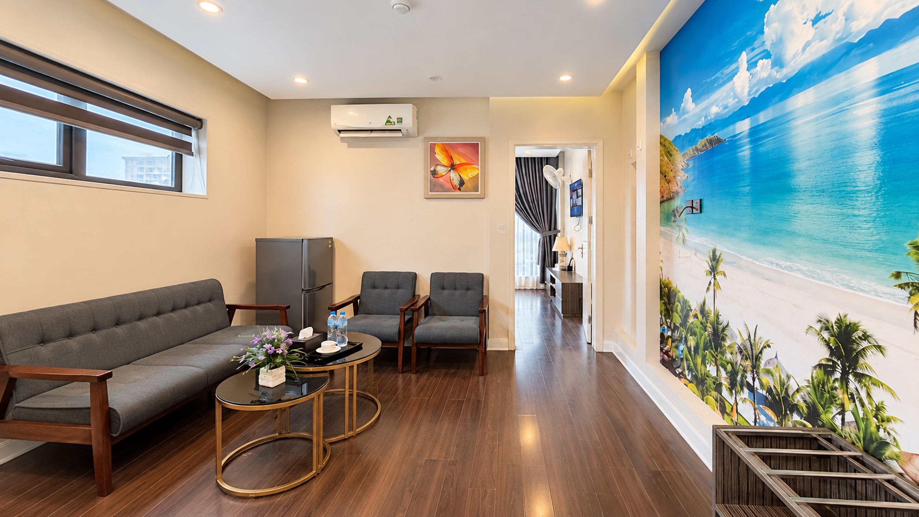 Ein modernes Wohnzimmer mit einer Couch, zwei Sesseln, einem Couchtisch und einem Kühlschrank. An der Wand befindet sich ein großes Wandbild mit Strandmotiv, und über der Couch ist eine Klimaanlage angebracht.