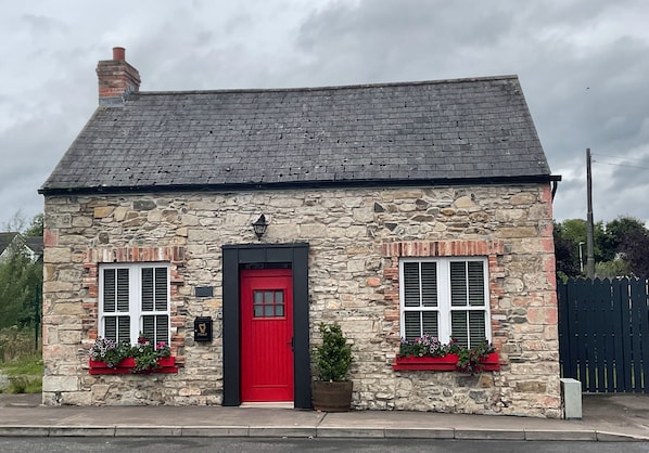 Un edificio de piedra con una puerta roja, dos ventanas con contraventanas y una chimenea.