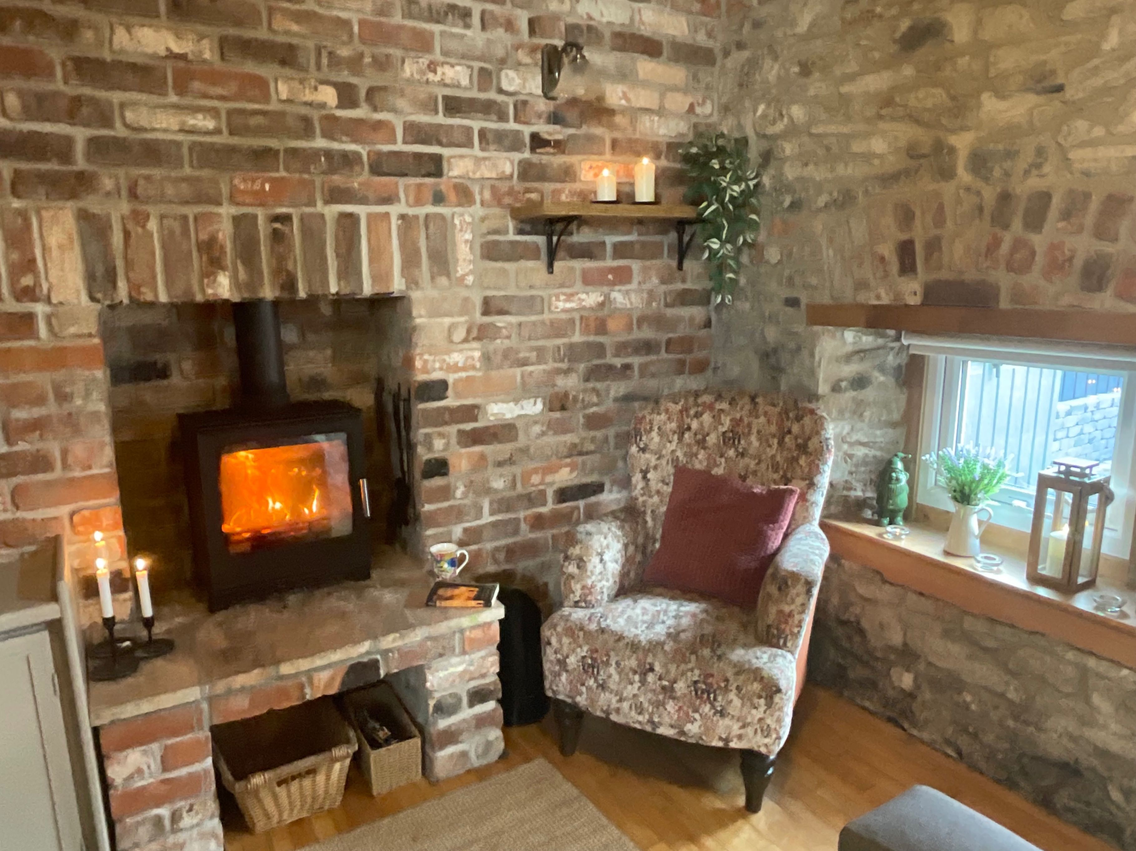 Una habitación acogedora con chimenea de ladrillo, un sillón floral y un estante con velas y plantas.