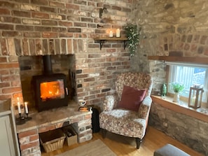 Una habitación acogedora con chimenea de ladrillo, un sillón floral y un estante con velas y plantas.