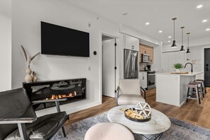 A modern living room with a fireplace, a flat-screen TV, and a dining area with stools. - Mountains Plenty | Luxury Hot Tub and Sauna (Canmore)