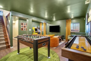 Une pièce comprenant une table de baby-foot, une table de jeu de palet, un canapé, un téléviseur et un escalier.