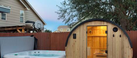 A wooden sauna structure with a bench inside, a hot tub outside, and a wooden fence in the background.