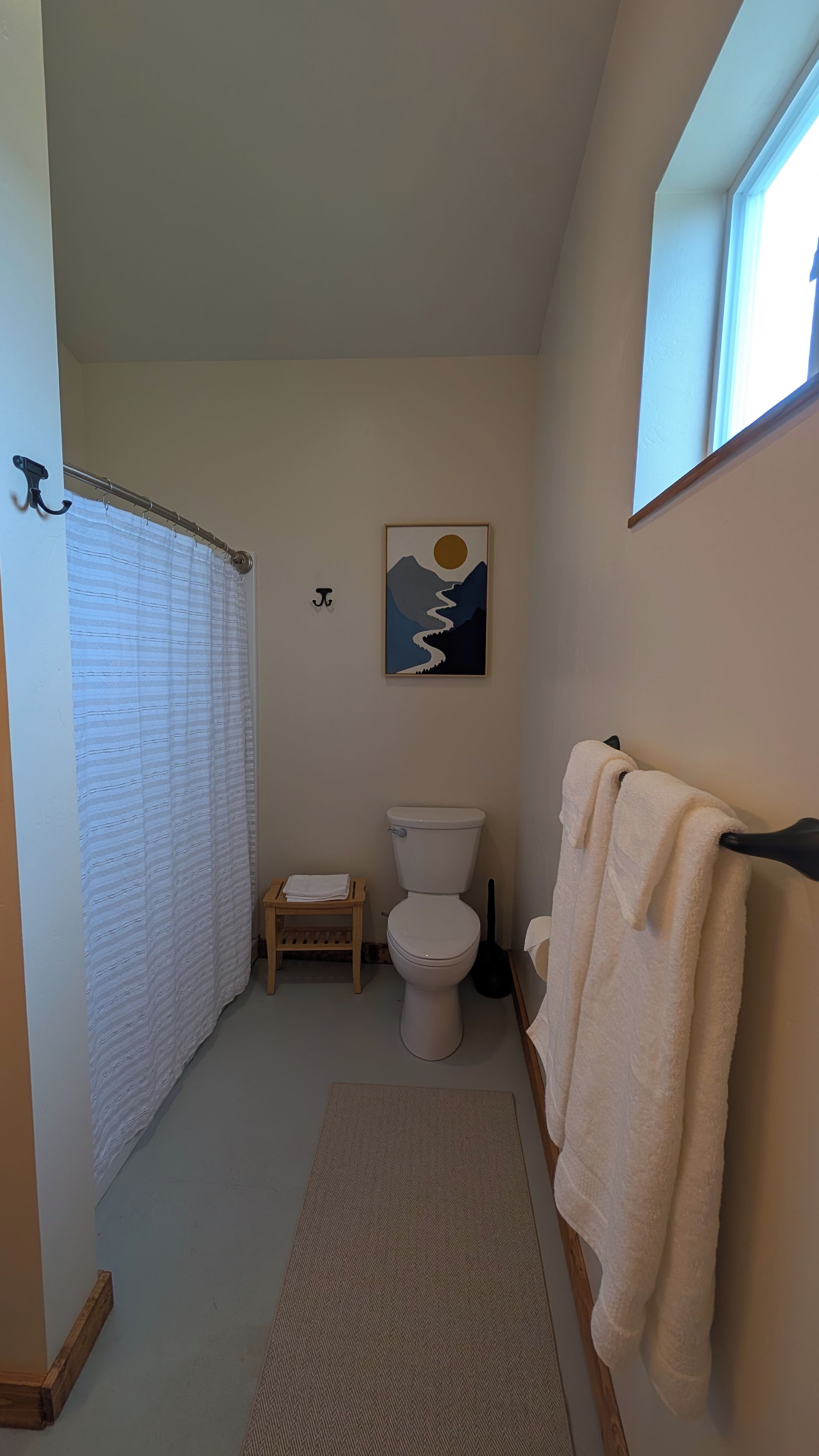 A bathroom with a toilet, a small wooden stool, a towel rack with towels, a painting on the wall, and a window.