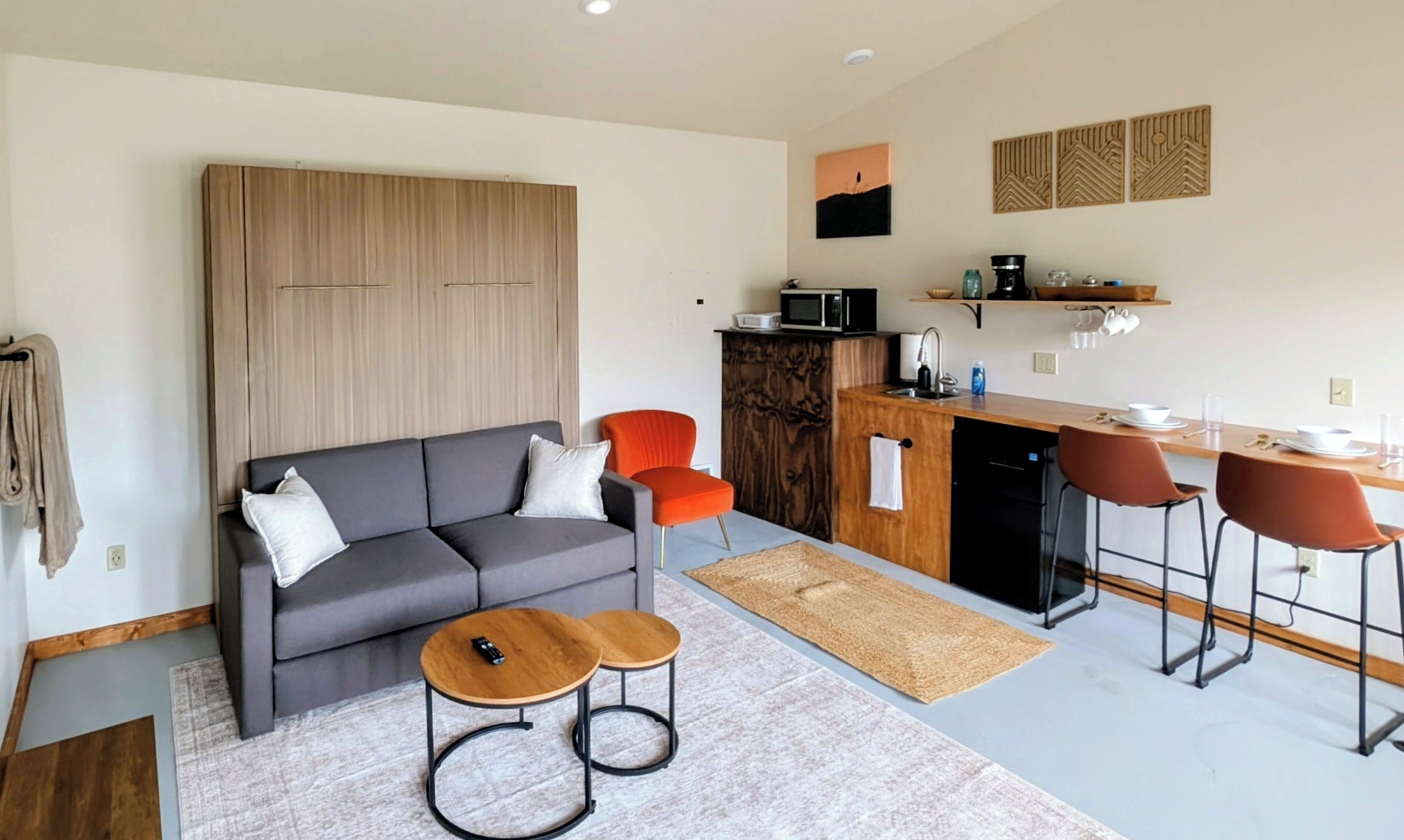 A modern living room with a grey sofa, two wooden side tables, and a kitchen area with a microwave, sink, and bar stools.