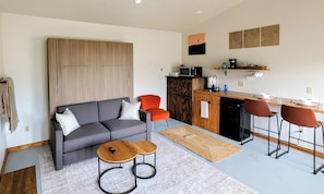 A modern living room with a grey sofa, two wooden side tables, and a kitchen area with a microwave, sink, and bar stools. - Amazing Views b/t Yellowstone and Livingston, Quiet & Private (Livingston)