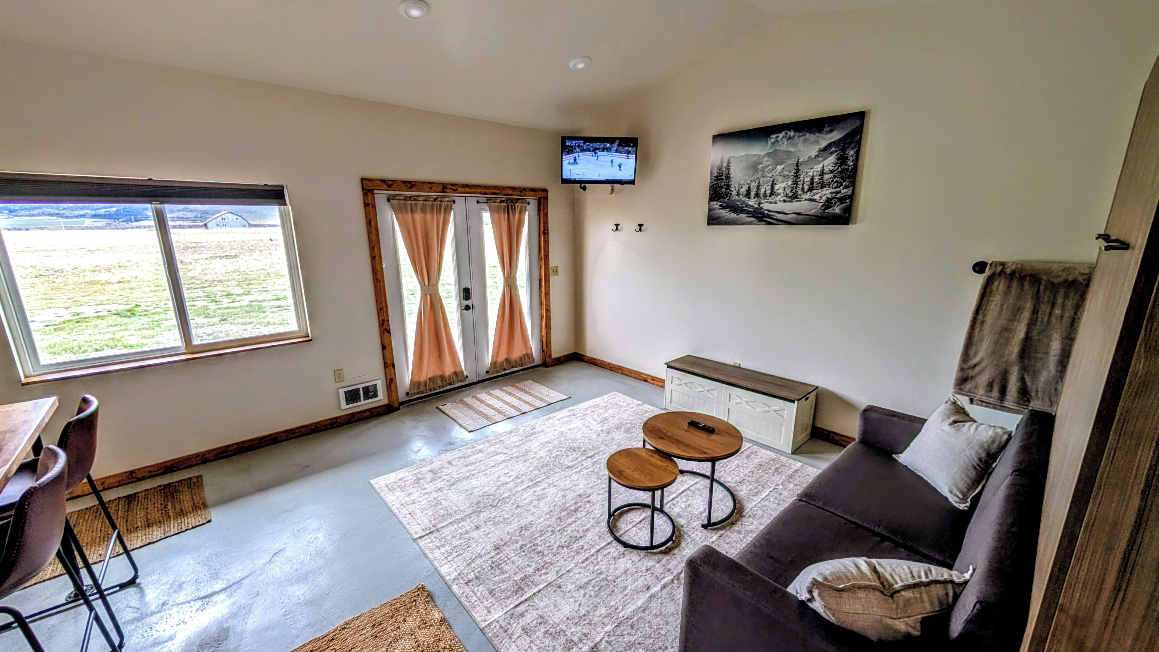 A living room with a sofa, two small tables, a TV mounted on the wall, and a window with curtains.