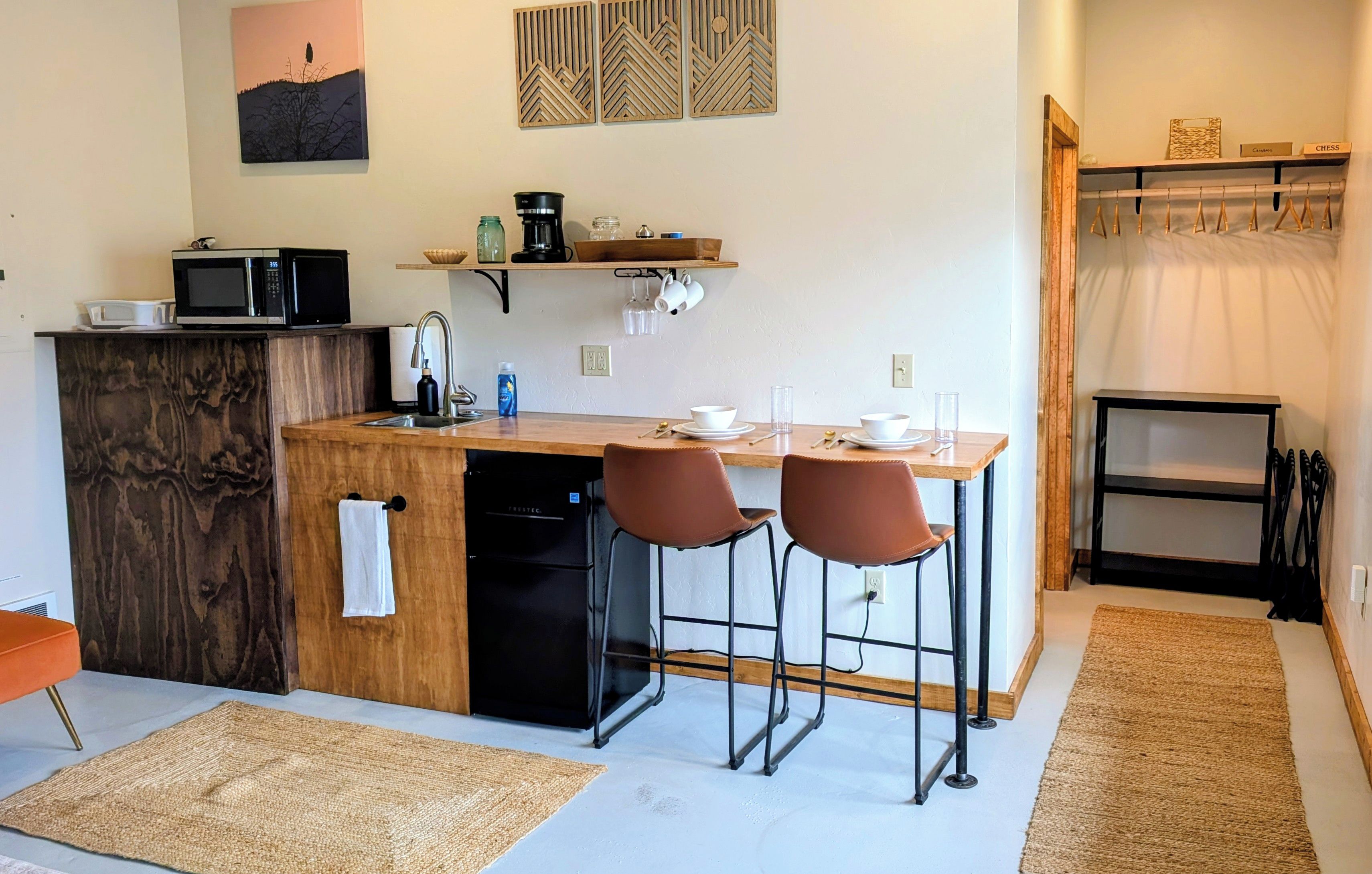 A compact kitchen with a microwave, coffee maker, and a sink. There are two bar stools and a small table with a few dishes.
