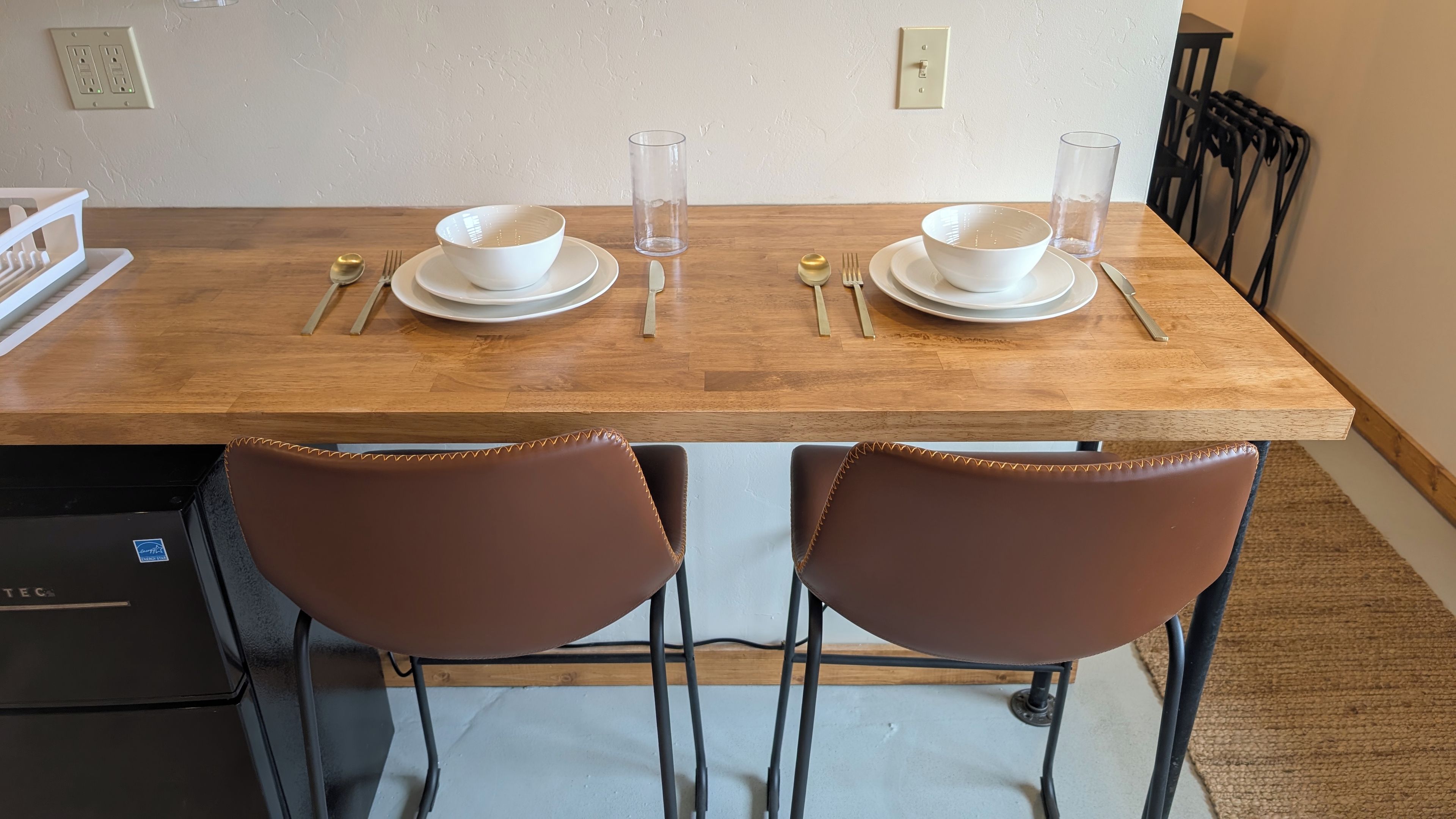 A dining table with two white bowls, a glass, and cutlery. There are two brown chairs with metal legs.