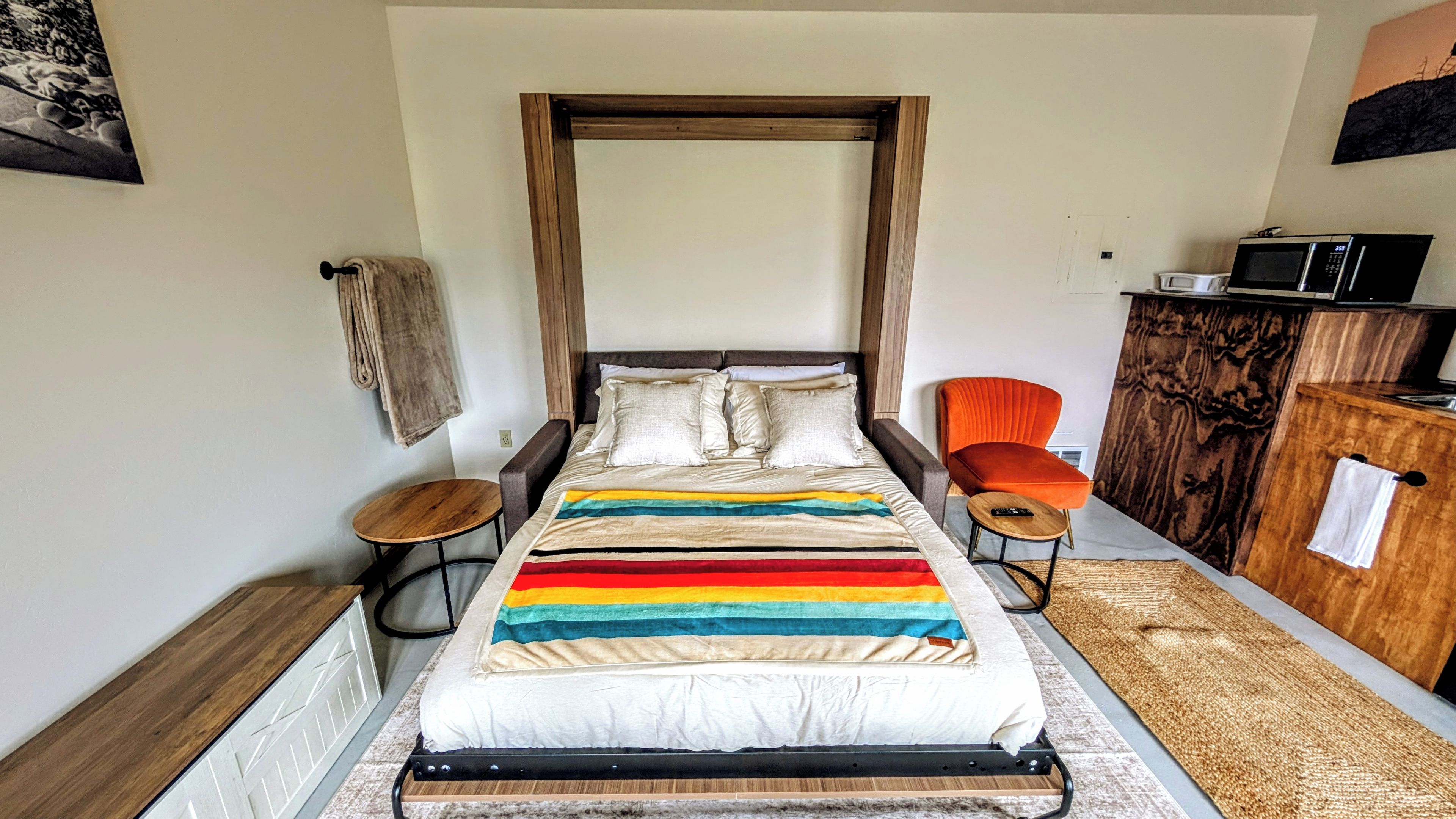 A bed with a rainbow-striped blanket, a wooden headboard, a small round table, a chair, and a microwave on a cabinet.
