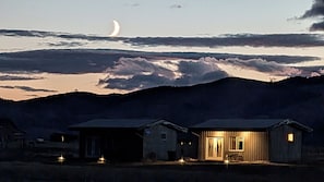 A two-story building with a porch and a lit entrance, surrounded by mountains and a crescent moon in the sky. - Amazing Views b/t Yellowstone and Livingston, Quiet & Private (Livingston)