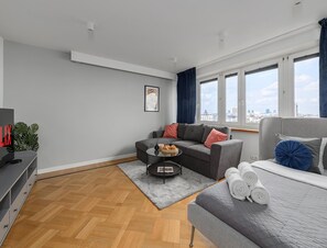 Una sala de estar moderna con un sofá gris, una mesita de centro, un televisor y vistas a la ciudad a través de amplias ventanas.
