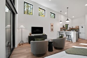 A modern living room with a flat-screen TV, two armchairs, a coffee table, and a kitchen area with a refrigerator, microwave, and bar stools. - Birchwood | Hocking Hills, Luxury, Hot Tub, Pets (McArthur)