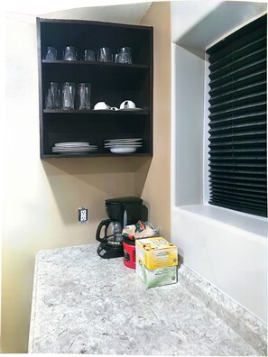 Una cocina con un estante negro que contiene cristalería y platos, una cafetera, una caja de té de limón y jengibre, y una caja de té de menta pura.