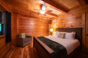 A wooden cabin room with a bed, a nightstand, a lamp, a window with curtains, and a ceiling fan. - Luna Moth | Hocking Hills, Firepit, Pool Table (Logan)