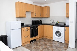 A compact kitchen with wooden cabinets, a white refrigerator, a microwave, an oven, a washing machine, and a sink. - Jacobs court 47 (Crawley)