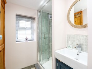 A bathroom with a glass-enclosed shower, a white sink, and a small window with blinds. - Beck Cottage (Sheringham)
