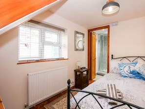 A bedroom with a bed, a radiator, a window with blinds, a bedside table with a lamp, and a door leading to a bathroom. - Beck Cottage (Sheringham)