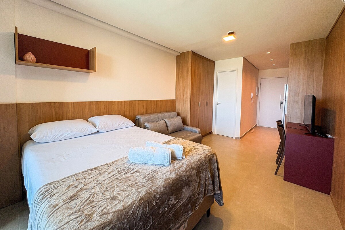 A hotel room with a large bed, a sofa, a desk with a television, and a door leading to another room.