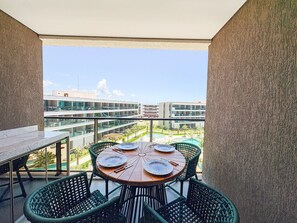 A balcony with a table set for four, surrounded by wicker chairs, overlooking a pool and buildings. - Beach Class Solare Muro Alto (Ipojuca)