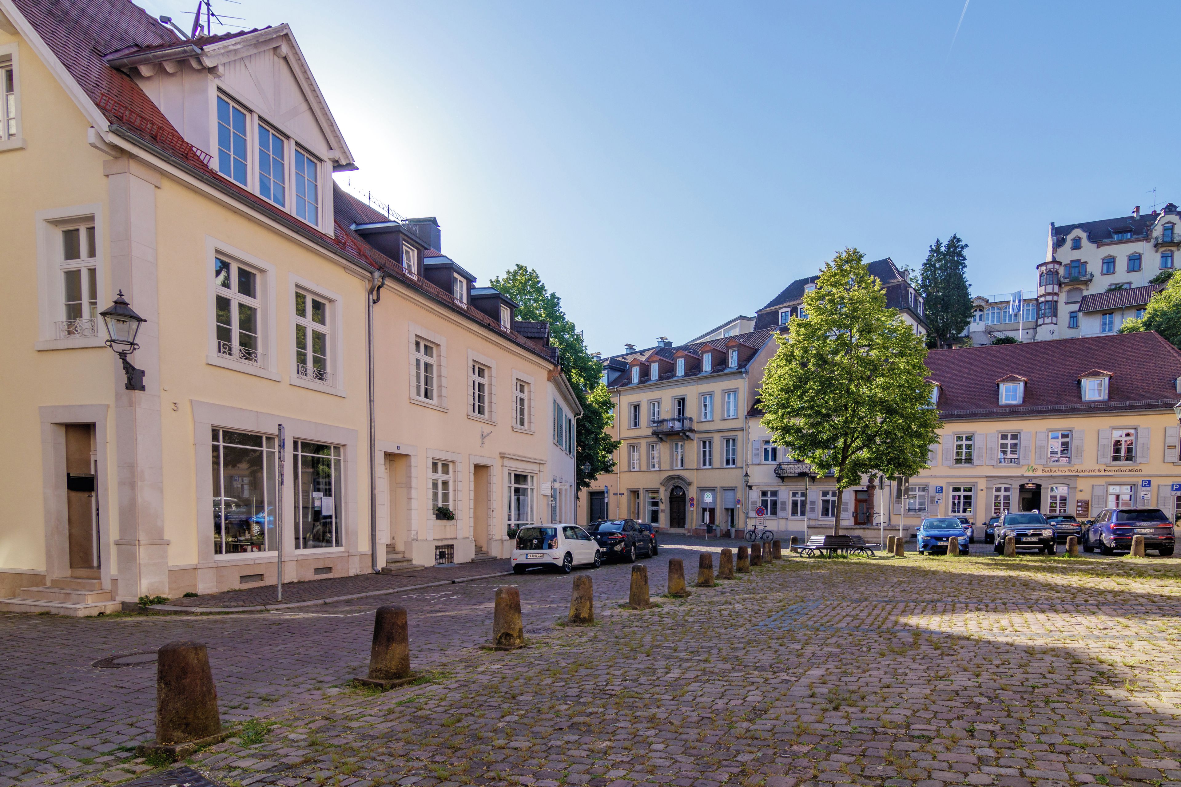 Eine Kopfsteinpflasterstraße gesäumt von historischen Gebäuden, einem Baum und parkenden Autos.