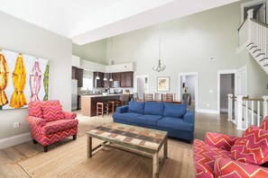 A spacious living area with a sofa, armchair, and coffee table. A kitchen is visible in the background with a dining area and a staircase. - 4BR Corolla Light Retreat w/ Hot Tub & Game Room (Corolla)