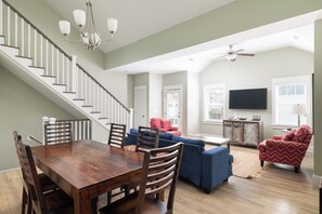 A spacious living area with a dining table, chairs, a blue sofa, a red patterned armchair, a ceiling fan, and a television mounted on the wall. - 4BR Corolla Light Retreat w/ Hot Tub & Game Room (Corolla)