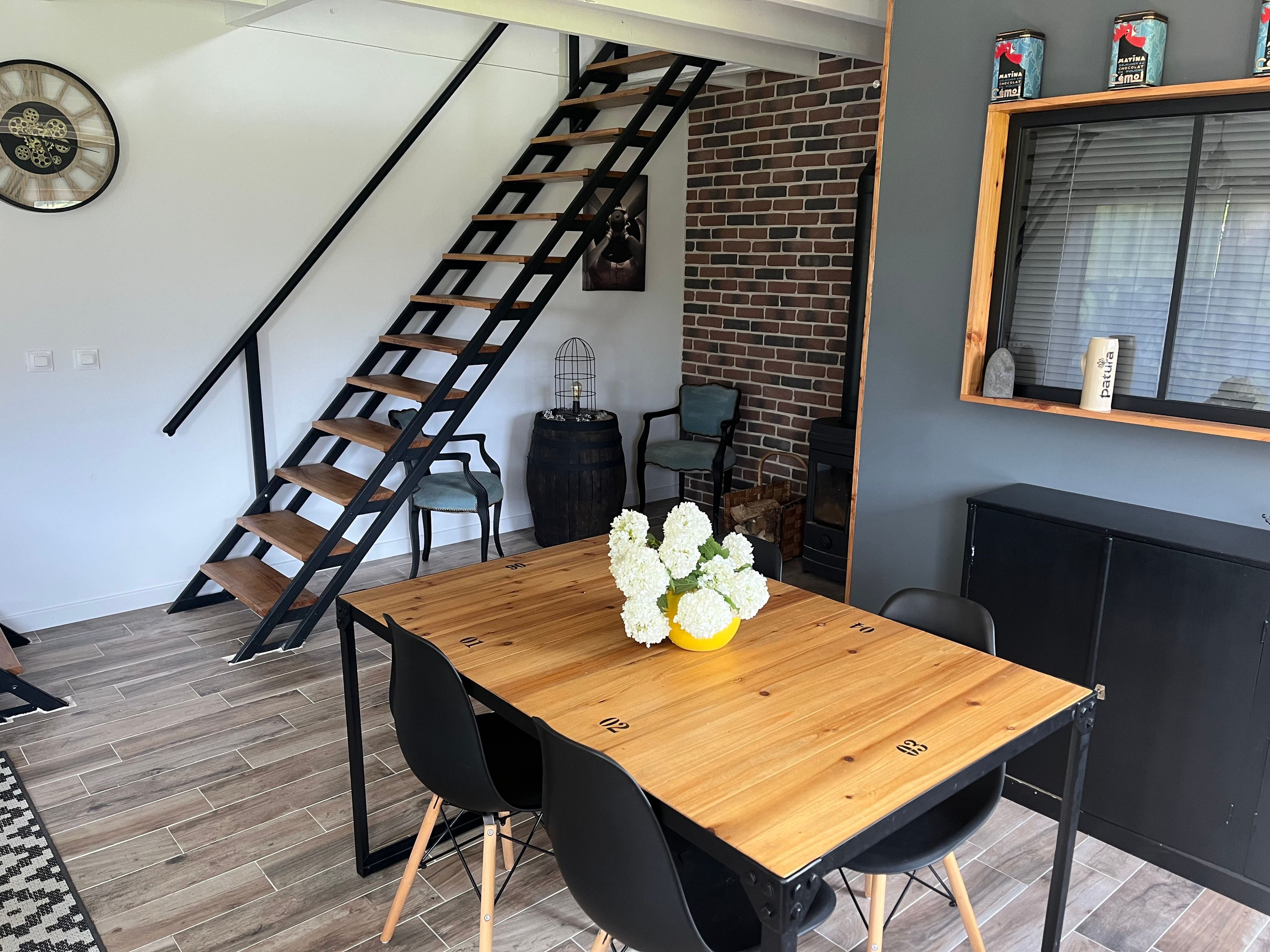 A modern interior with a wooden dining table, black chairs, a brick wall, a black staircase, and a large window.