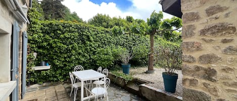 A stone patio with a white table and chairs, surrounded by a stone wall and a hedge.