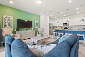 Uma sala de estar moderna com uma parede de destaque verde, uma televisão de tela plana grande e um sofá com almofadas azuis.