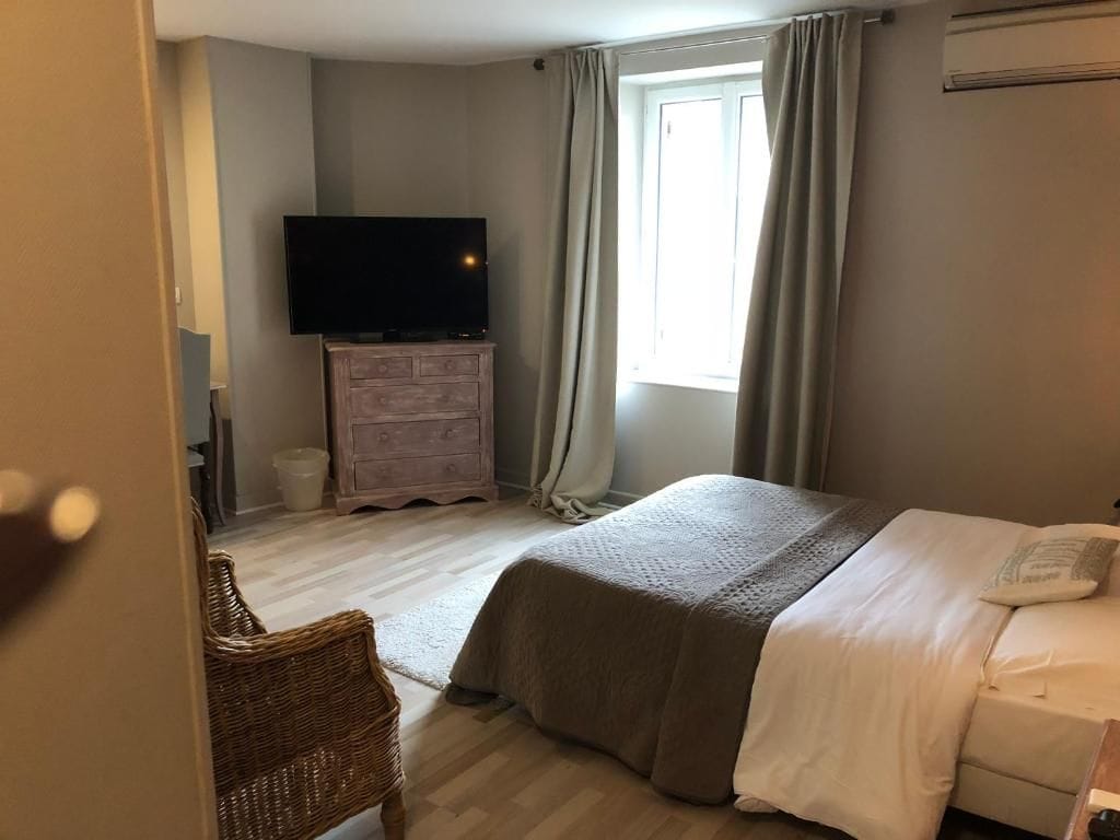 A bedroom with a bed, a television on a dresser, a window with curtains, and a wicker chair.