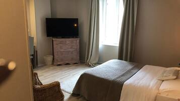 A bedroom with a bed, a television on a dresser, a window with curtains, and a wicker chair.