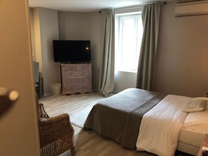 A bedroom with a bed, a television on a dresser, a window with curtains, and a wicker chair. - LE CHENE PENDRAGON (Saint-Léger-en-Yvelines)