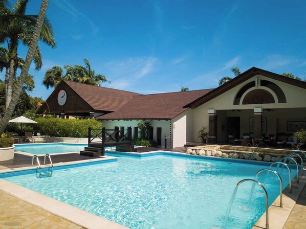 A swimming pool with a sunshade, a covered seating area, and a building with a clock. - Junior Suite – Beachfront Escape on Cofresí Beach (Puerto Plata)