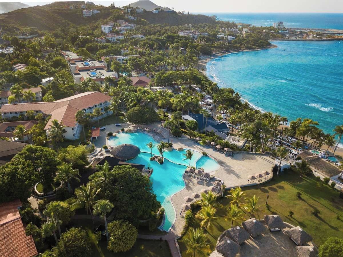 A resort with a large swimming pool, palm trees, and a beach area.