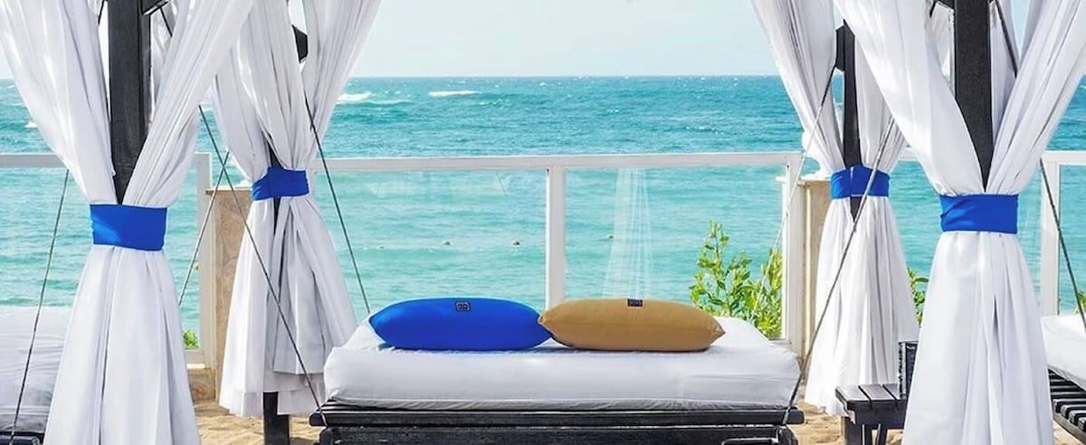 A bed with white bedding and two pillows, a white canopy with a blue ribbon, and a view of the ocean.