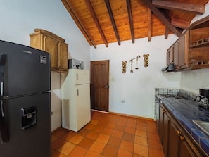 Una cocina con un refrigerador negro, un refrigerador blanco, armarios de madera, piso de baldosas y techo de madera.