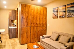 A living room with a sofa, a wooden wardrobe, a kitchen area, and a wall with paintings. - Estudio Moderno Caribe  o  con Vista al Mar (Playa del Carmen)