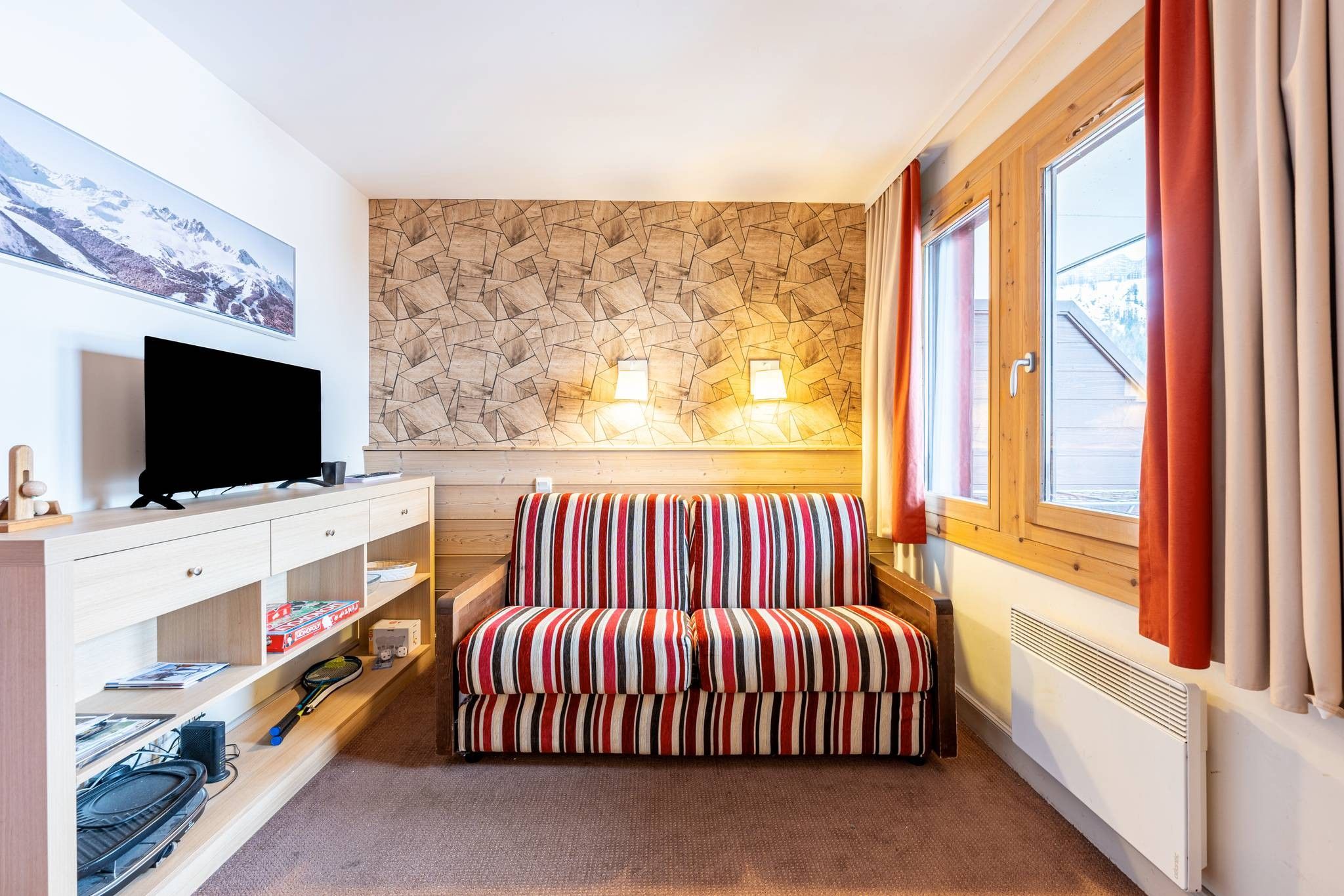 Une chambre d’hôtel avec un canapé à rayures, un téléviseur à écran plat, un mur en bois orné de motifs géométriques, deux lampes fixées au mur et une fenêtre avec des rideaux.