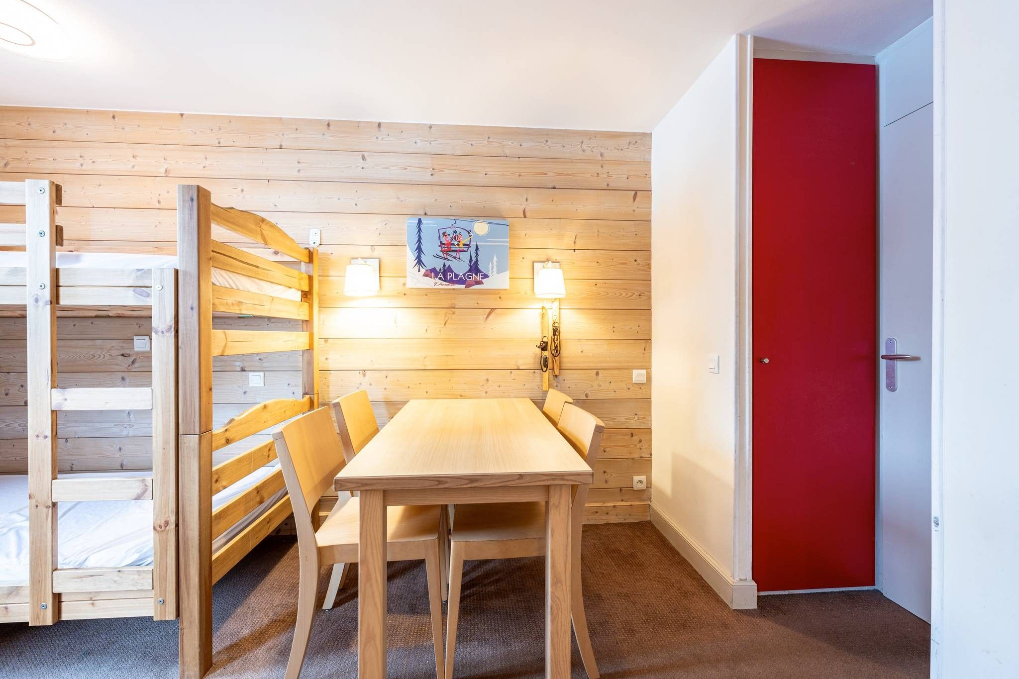 Une pièce avec un lit superposé en bois, une table avec des chaises, une porte rouge et un luminaire fixé au mur.