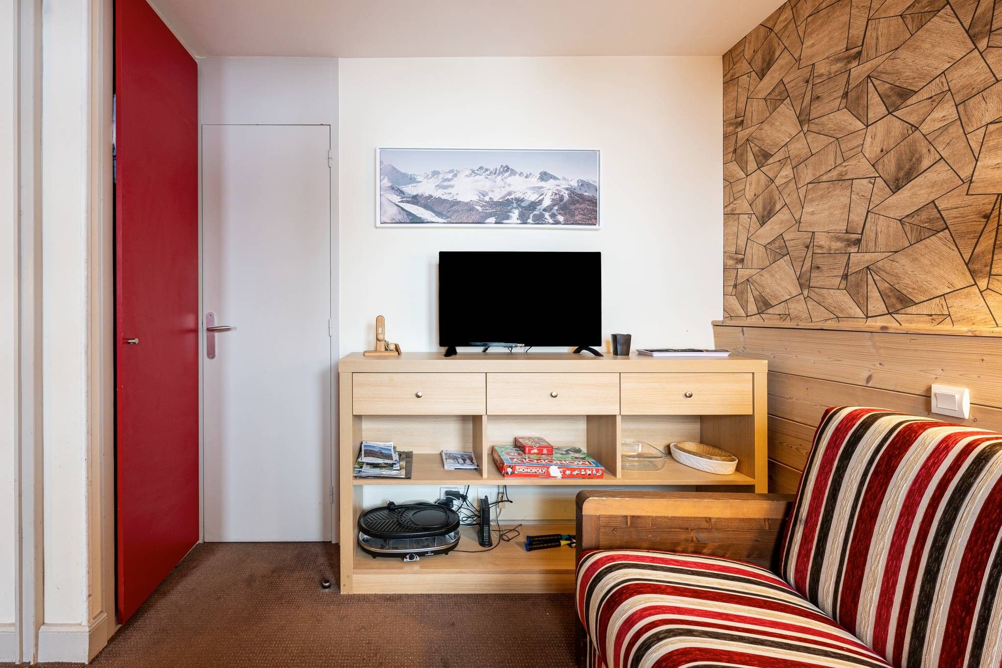 A compact living space with a red door, a television on a wooden stand, a striped sofa, and a wall-mounted picture.