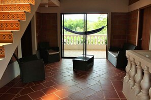Una habitación con suelo de baldosas, una hamaca, muebles de mimbre y un balcón con barandilla blanca.