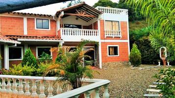 A two-story house with a balcony, surrounded by a gravel driveway and lush greenery.