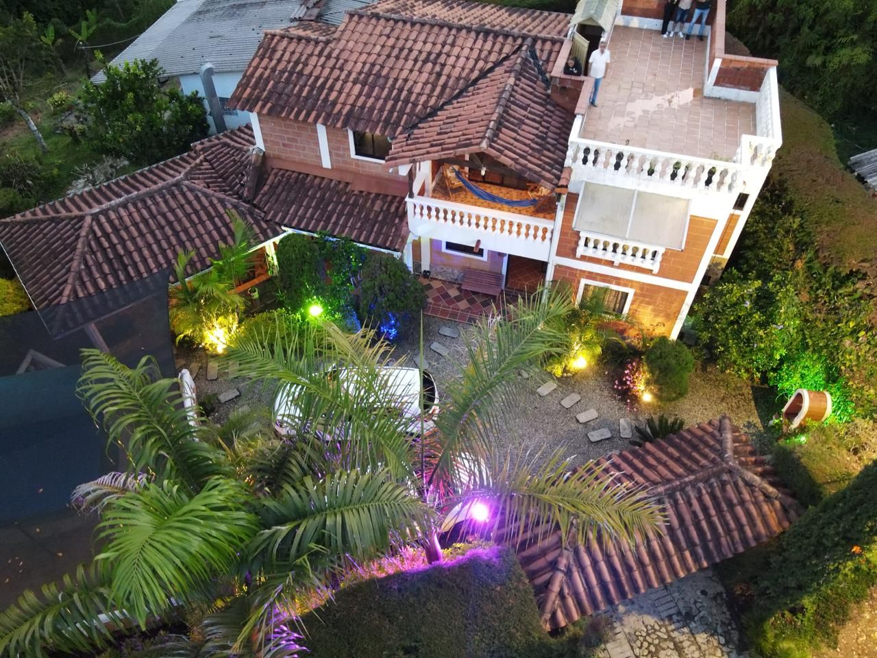 Un edificio de dos pisos con techo de tejas, balcón y un espacio exterior bien iluminado con plantas e iluminación decorativa.