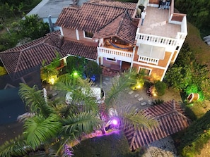 Un edificio de dos pisos con techo de tejas, balcón y un espacio exterior bien iluminado con plantas e iluminación decorativa.