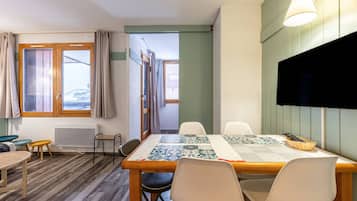 A cozy room with a wooden dining table, four chairs, a TV mounted on the wall, and a window with curtains.