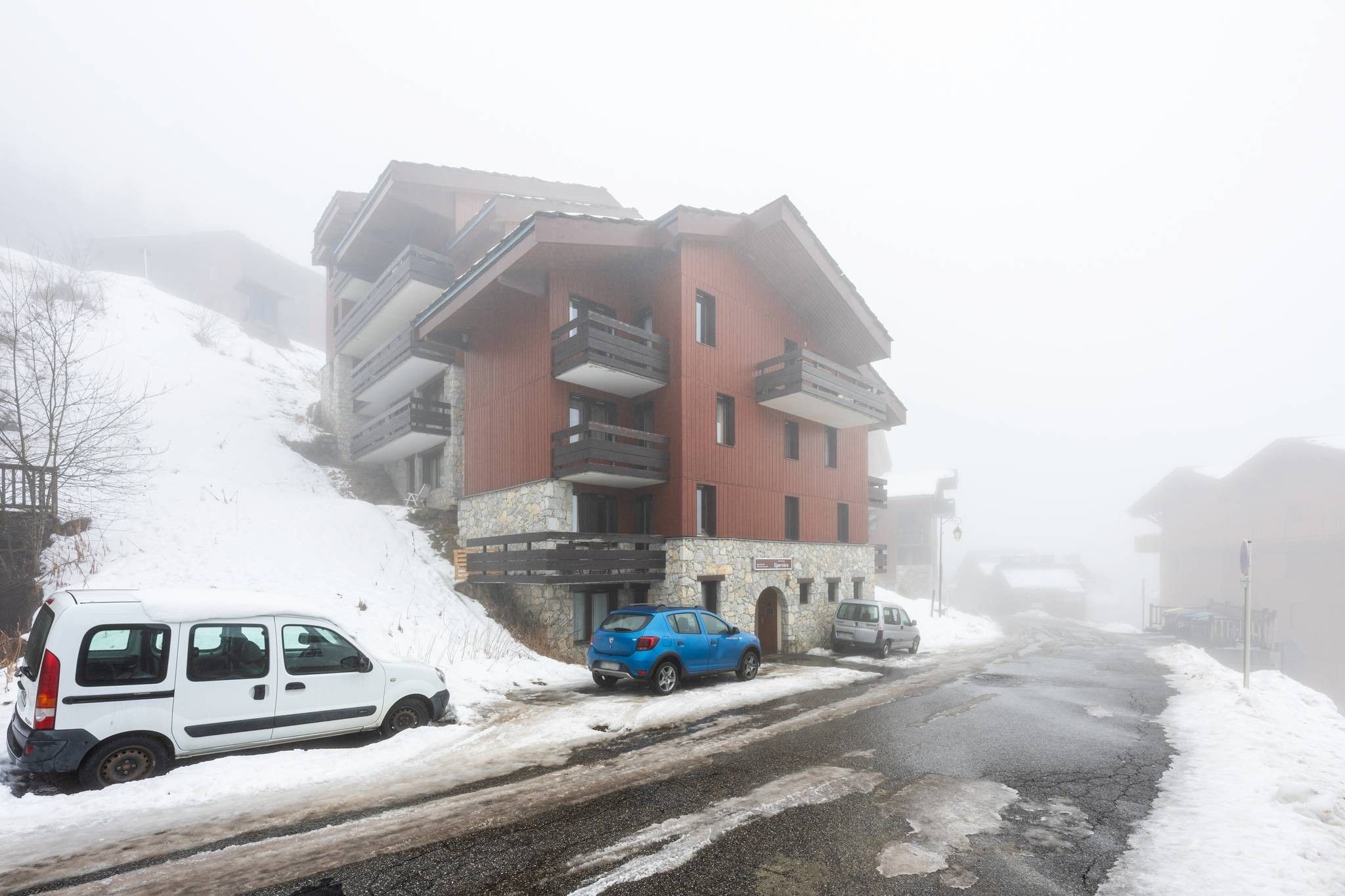 A multi-story building with balconies, a snow-covered street, and parked vehicles.